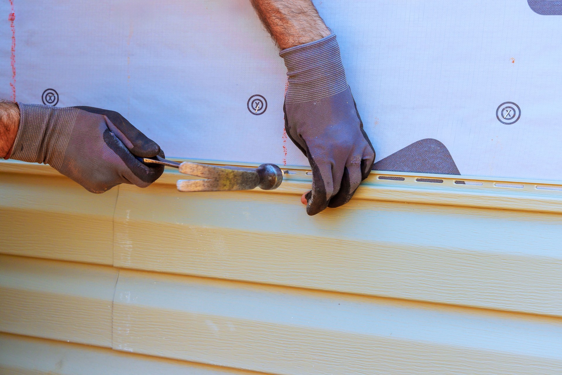 Worker installs siding on a house using a hammer and level during a sunny day in a residential area